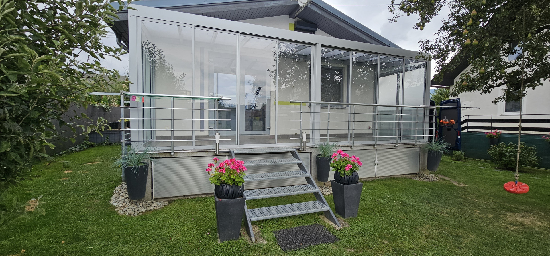 Une véranda moderne vitrée, située sur une terrasse surélevée, avec un escalier et des compositions florales