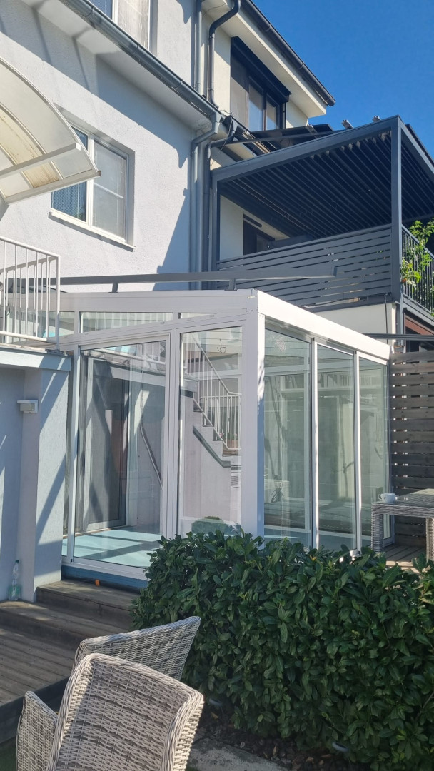 Une véranda moderne, blanche et vitrée, attenante à une maison individuelle, avec vue sur la terrasse et la verdure.