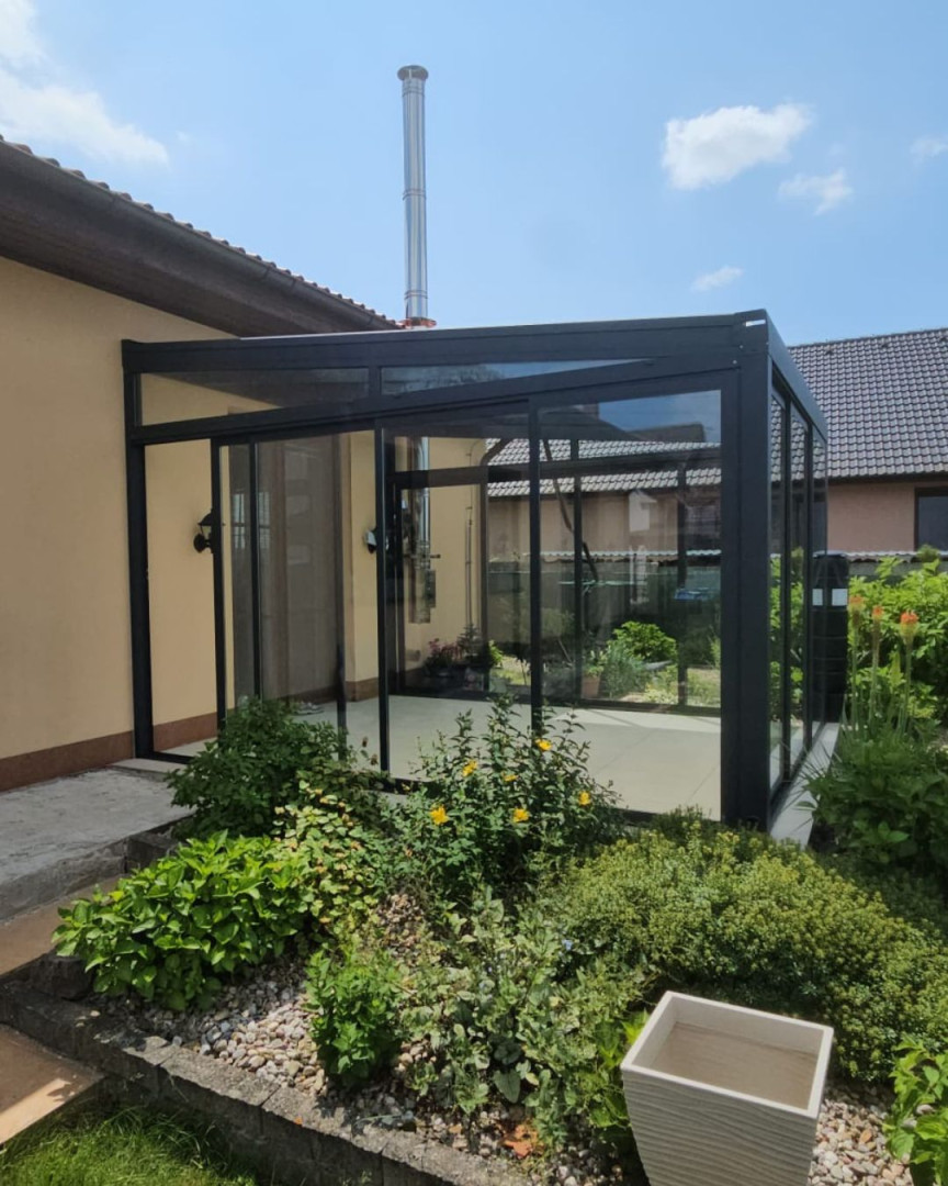 Une extension moderne vitrée adossée à une maison individuelle, dotée d'un cadre noir et de portes coulissantes, entourée d'un jardin.