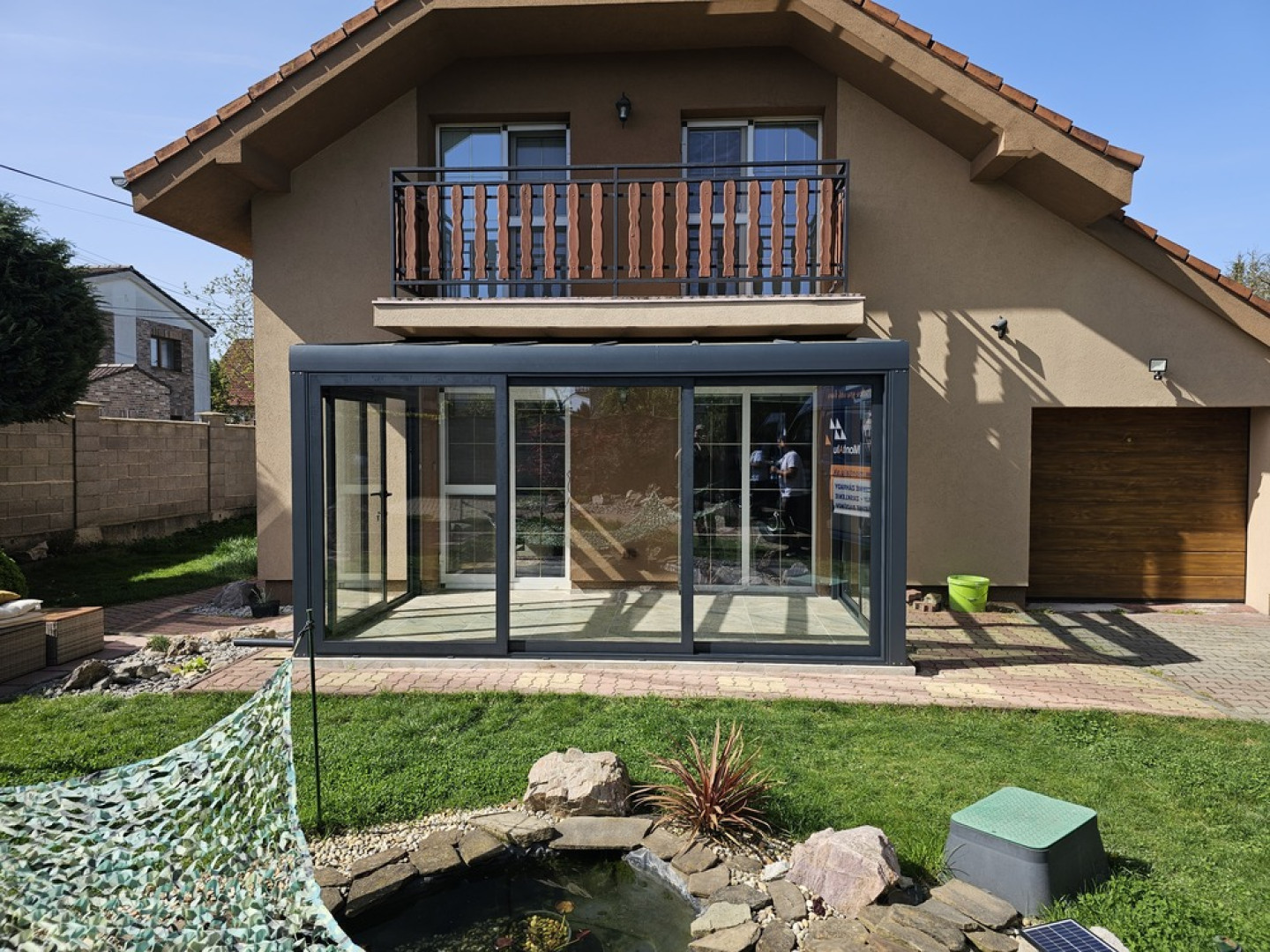 Une véranda moderne vitrée, dotée d'une structure anthracite, adossée à une maison individuelle, avec un petit étang de jardin au premier plan.