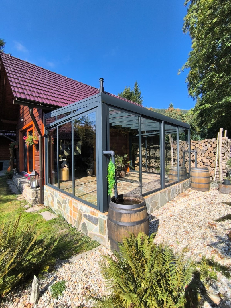 Une véranda moderne vitrée, dotée d'un cadre en aluminium foncé, adossée à une maison en bois avec un socle en pierre.
