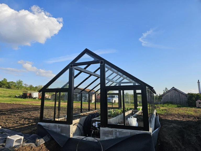 Une serre moderne vitrée, dotée d'une structure noire, construite sur des fondations en béton dans un jardin sous un ciel bleu.