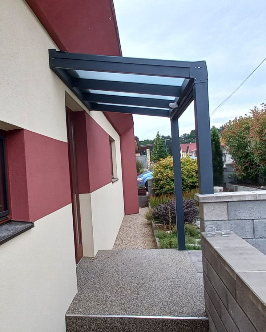Une pergola moderne en aluminium gris foncé avec un toit en verre, installée au-dessus de l'entrée de la maison.