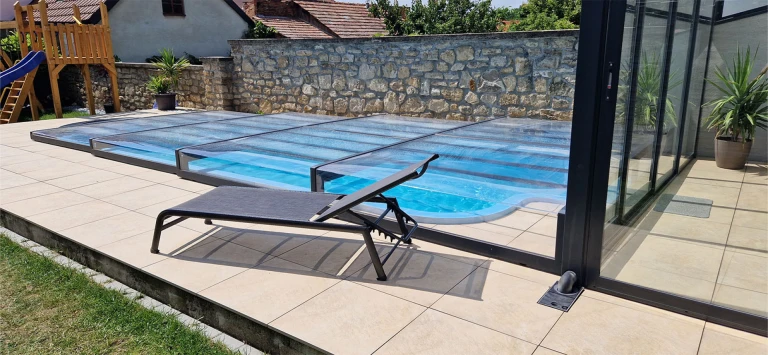 Un abri de piscine moderne et bas, doté d'une structure en aluminium noir, installé sur une terrasse équipée d'une chaise longue
