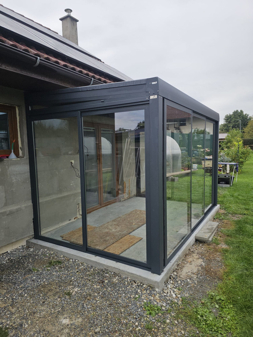 Une extension moderne vitrée servant de jardin d'hiver, dotée d'une structure gris foncé, attenante à une maison individuelle